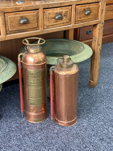 A vintage copper fire extinguisher with a brass handle and lid, featuring an embossed brass label on the front